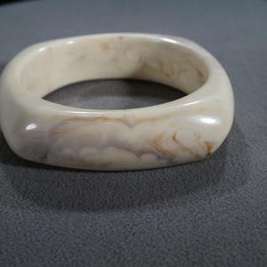 May include: A white and brown marbled plastic bangle bracelet. The bracelet is square shaped and has a smooth, glossy finish.
