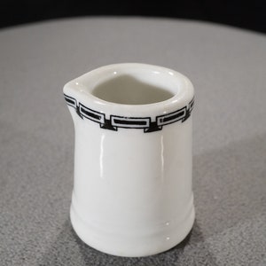 May include: A small white ceramic creamer with a black and white geometric design around the top rim.