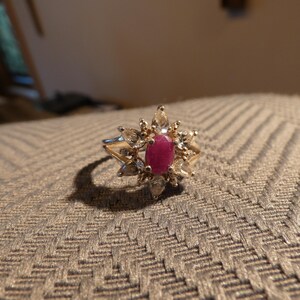 May include: A gold ring featuring a central oval ruby gemstone, surrounded by smaller, clear gemstones in a starburst design. The ring is set on a textured surface.