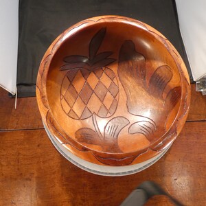 May include: A wooden bowl with a carved pineapple design. The bowl is made of a light brown wood and has a round shape. The pineapple is carved in the center of the bowl, with leaves and other decorative elements around it.