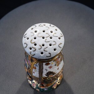 May include: A white ceramic salt shaker with a gold trim and a floral design. The top of the shaker has a circular pattern of holes and gold accents.