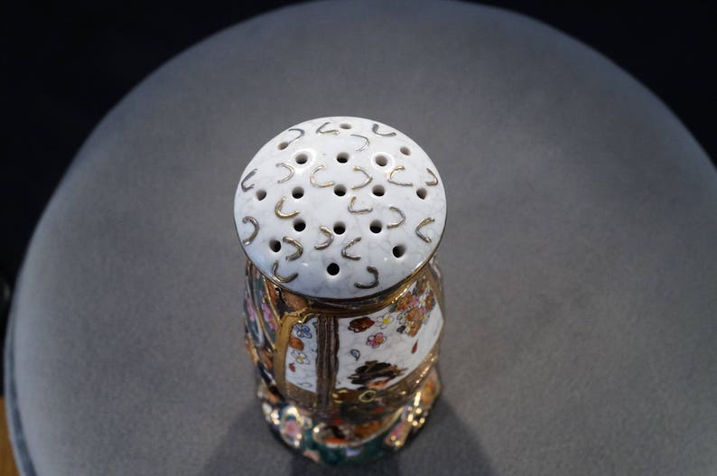 May include: A white ceramic salt shaker with a gold trim and a floral design. The top of the shaker has a circular pattern of holes and gold accents.
