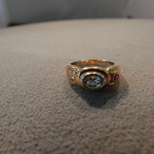 May include: A gold ring featuring a central oval-cut light blue stone in a bezel setting. The band includes small clear accent stones on one side and a single round red stone on the other side, with a polished gold finish throughout the design.