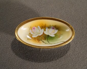 May include: A small, oval-shaped dish with a gold rim. The dish is decorated with a floral design featuring two white roses with pink centers, set against a yellow and green background.