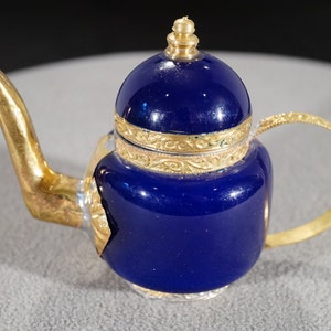 May include: A small blue teapot with gold accents and a lid. The teapot has a gold spout and handle.