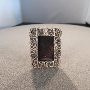 May include: A rectangular silver-toned picture frame with an ornate, scrolling design around the edges. The frame has a dark, possibly black, interior. The frame is likely made of metal and is designed to hold a small photograph or artwork.