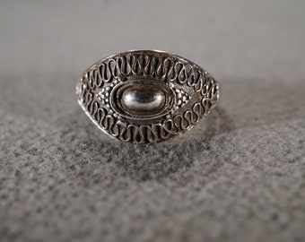 May include: A silver ring with an oval-shaped center stone surrounded by a decorative border of swirling designs.