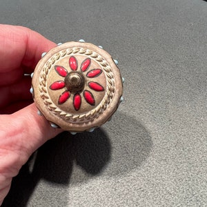 May include: A round, beige ceramic knob with a raised, gold-colored rope design around the edge. The knob has a red and white flower design in the center.