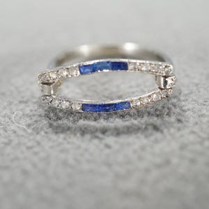 May include: A silver ring with a unique design. The ring features a curved band with small, clear stones and two rectangular blue stones. The ring is set against a gray textured background.