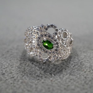May include: A silver-toned ring with a central oval-shaped green gemstone, surrounded by smaller clear stones. The ring has an ornate, openwork design with a cluster of stones around the central gem.