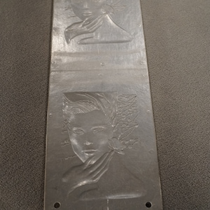 May include: A silver metal plate with two embossed images of a woman's face. The woman's face is surrounded by leaves and flowers. The plate has a textured surface and is slightly curved.