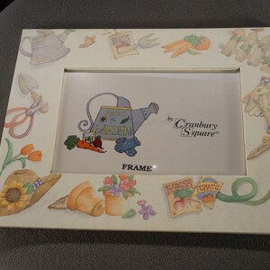 May include: White picture frame with a garden theme. The frame has a light blue watering can with the words "Drink in the Garden" on it. The frame also has a variety of other garden-themed images, including carrots, radishes, tomatoes, and gardening gloves. The text "by Cranberry Square" is printed on the frame.