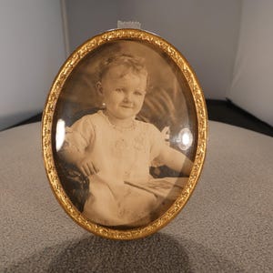 May include: An oval-shaped gold-toned metal picture frame with an ornate, textured border. The frame contains a vintage black and white photograph of a young child wearing a light-colored dress with ruffled sleeves and a delicate collar detail.