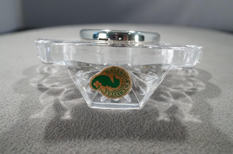 May include: A clear glass clock base with a silver clock face and a green and white sticker that reads "WATERFORD CRYSTAL".