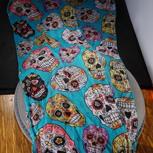 May include: A turquoise blue scarf with a repeating pattern of colourful sugar skulls. The skulls are decorated with flowers and other intricate designs.