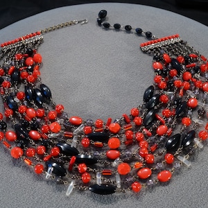 May include: Multi-strand necklace with black and red beads. The necklace is made of several strands of beads, including black oval beads, red round beads, and clear beads. The necklace is secured with a silver clasp.