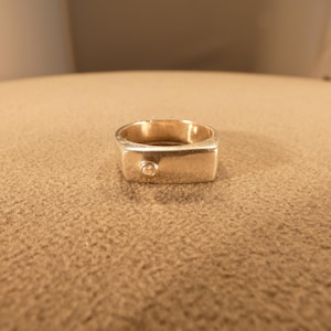 Puede incluir: Un anillo dorado con una cara rectangular y una pequeña piedra redonda y transparente incrustada en la cara. El anillo está sobre un fondo neutro y texturizado.