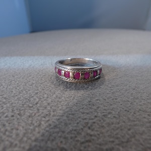 May include: A silver-toned band ring featuring a central row of small, round, vibrant pink stones set between two decorative twisted rope-style borders. The metal has a polished finish, and the design creates a repeating pattern around the band.