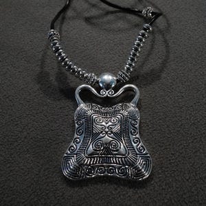 May include: A silver necklace with a black leather cord and a silver pendant with an intricate design. The pendant is shaped like a shield with a square in the center and a swirl pattern around the edges.