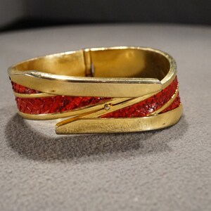 May include: A gold-toned hinged bangle bracelet with red leather inlay. The bracelet has a geometric design and is accented with a small rhinestone.