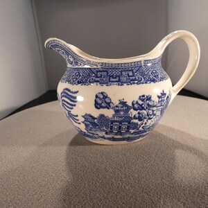 May include: Cream-colored creamer with a blue Willow pattern design. The creamer has a curved spout and a handle. The blue pattern features a landscape with buildings, trees, and a fence. The creamer is made of ceramic.