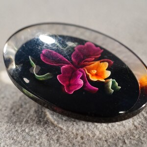 K&ouml;nnte beinhalten: Ein ovaler Knopf aus klarem Glas mit schwarzem Hintergrund und einem bunten Blumendesign. Die Blume ist rosa, orange und gr&uuml;n.