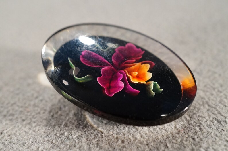 K&ouml;nnte beinhalten: Ein ovaler Knopf aus klarem Glas mit schwarzem Hintergrund und einem bunten Blumendesign. Die Blume ist rosa, orange und gr&uuml;n.