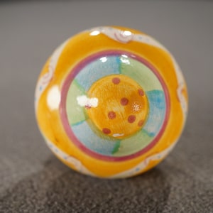 May include: A yellow ceramic ornament with a multi-colored design. The ornament features a central circle with a yellow background and red polka dots, surrounded by concentric circles of green, blue, pink, and white.