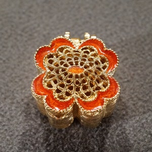 May include: A gold-colored metal trinket box shaped like a flower with orange enamel accents.