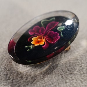 K&ouml;nnte beinhalten: Eine transparente ovale Brosche mit schwarzem Hintergrund und einem farbenfrohen Blumendesign. Die Blume ist rosa, gelb und orange mit gr&uuml;nen Bl&auml;ttern.