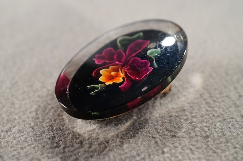 K&ouml;nnte beinhalten: Eine transparente ovale Brosche mit schwarzem Hintergrund und einem farbenfrohen Blumendesign. Die Blume ist rosa, gelb und orange mit gr&uuml;nen Bl&auml;ttern.