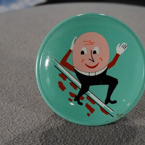 May include: A vintage tin plate with a turquoise blue background featuring a cartoon character with a large pink head and black body, skiing down a red brick wall.