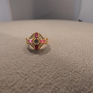 May include: A gold-toned ring with a central diamond-shaped design. The ring features a central gray stone, surrounded by pink and clear stones. The ring is set on a neutral-colored surface.