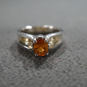 May include: A silver and gold ring with an oval, orange gemstone. The ring has a gold band and a silver setting around the gemstone. The ring is set against a gray background.