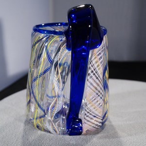 May include: A clear glass mug with a blue handle. The mug has a swirling pattern of white, yellow, and blue lines.