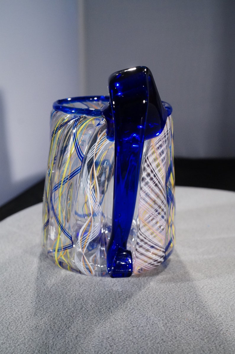 May include: A clear glass mug with a blue handle. The mug has a swirling pattern of white, yellow, and blue lines.
