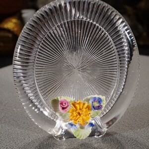 May include: A clear glass coaster with a raised, textured design. The coaster has a floral motif with a pink rose, yellow carnation, and blue pansy.