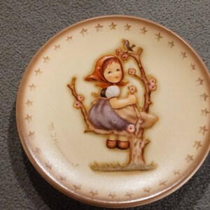 May include: A round, cream-colored plate with a brown border and a white star pattern. The center features a girl in a red and white dress, sitting on a branch with pink flowers and a bird.