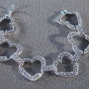 May include: A silver bracelet with a heart-shaped link design. The bracelet is made of metal and has a shiny finish. The hearts are decorated with small, black stones.