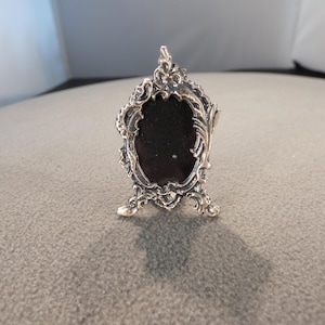 May include: Ornate silver-toned picture frame with a dark interior. The frame has an oval shape with decorative scrollwork and stands on two small feet. A small, elegant decorative item.