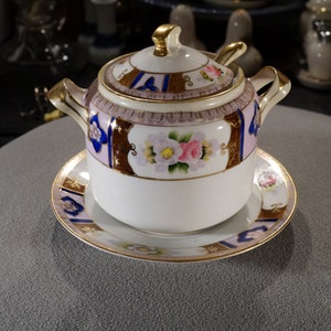 May include: A white porcelain sugar bowl with a lid and a matching saucer. The bowl and saucer are decorated with gold trim and floral designs in shades of pink, blue, and brown.