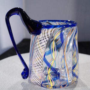 May include: A clear glass mug with a blue handle and colorful swirls of glass throughout the body.