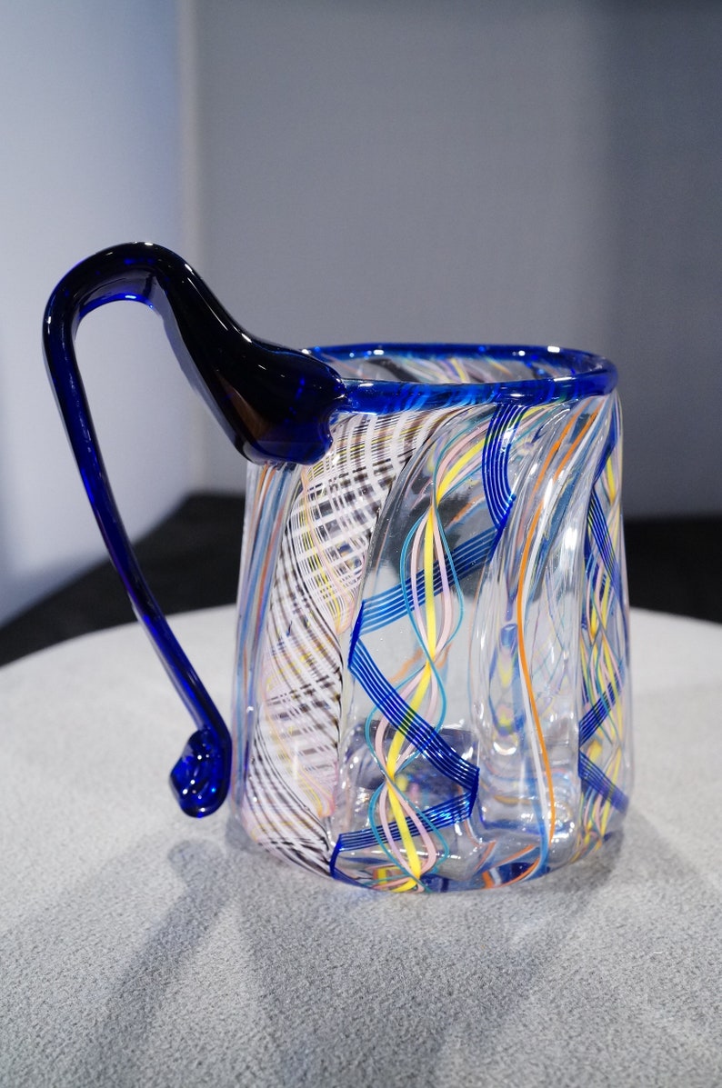 May include: A clear glass mug with a blue handle and colorful swirls of glass throughout the body.
