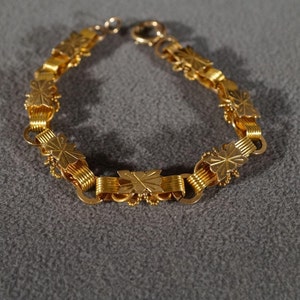 May include: A gold bracelet with a chain link design featuring a repeating pattern of stylized leaves. The bracelet has a simple clasp.