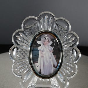 May include: A clear crystal picture frame with a scalloped edge and a silver oval frame. The frame features a picture of a young girl wearing a white dress and a crown, holding a wand.
