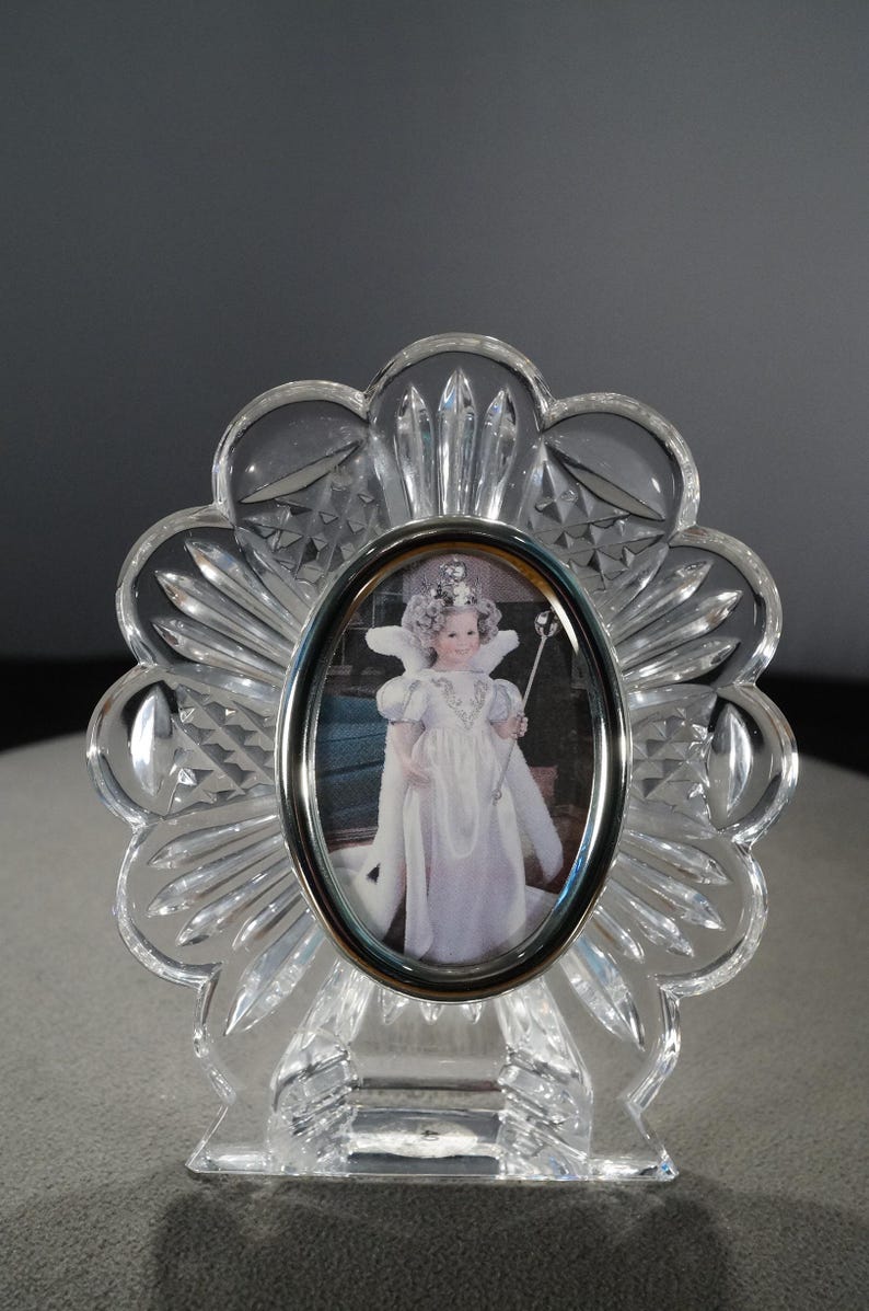 May include: A clear crystal picture frame with a scalloped edge and a silver oval frame. The frame features a picture of a young girl wearing a white dress and a crown, holding a wand.