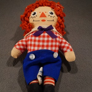 May include: A red-haired rag doll with a red and white checkered shirt, blue pants, and black shoes. The doll has a purple bow and a painted face with black eyes and a red mouth.