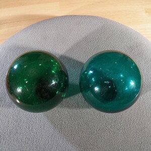 May include: Two green glass balls, one slightly darker than the other, are sitting on a gray surface.