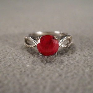May include: A silver ring with a large, round, red gemstone set in the center. The band is decorated with smaller, white gemstones.
