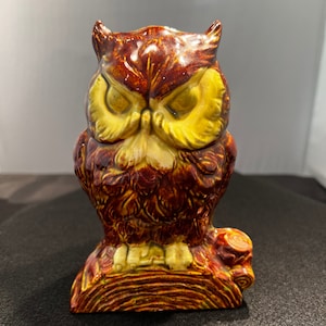 May include: A ceramic owl figurine with brown and yellow glaze, perched on a tree stump. The owl has large, round eyes and a detailed feather pattern.